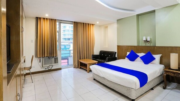 Ein Hotelzimmer mit einem großen Bett, einem Schreibtisch, einem Stuhl, einem Fernseher und einem Balkon mit Vorhängen.