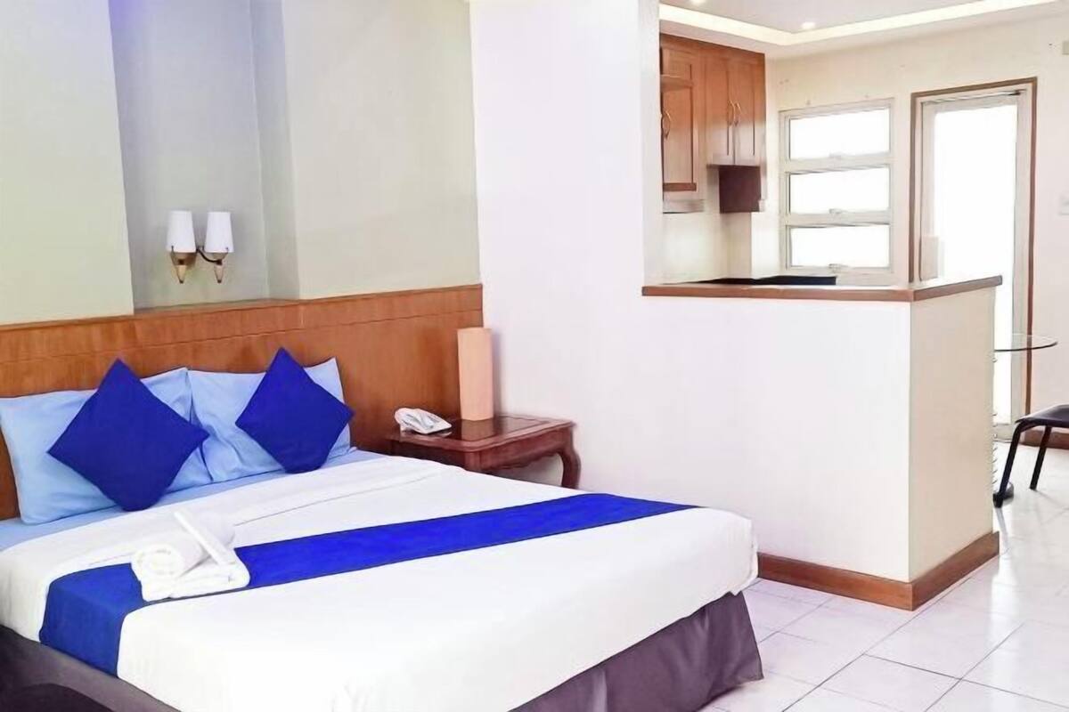 A hotel room with a bed, a bedside table with a phone, a small kitchenette, and a door leading to a bathroom.