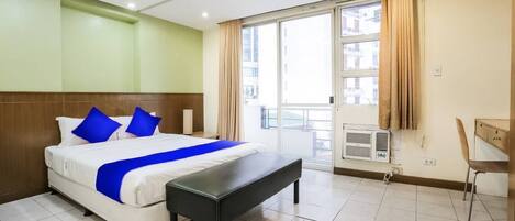 A hotel room with a bed, a bench, a desk, and a window with curtains.