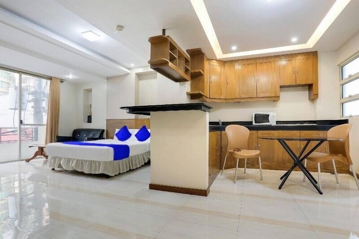A modern hotel room with a bed, a desk, a chair, a small kitchen area, and a large window.