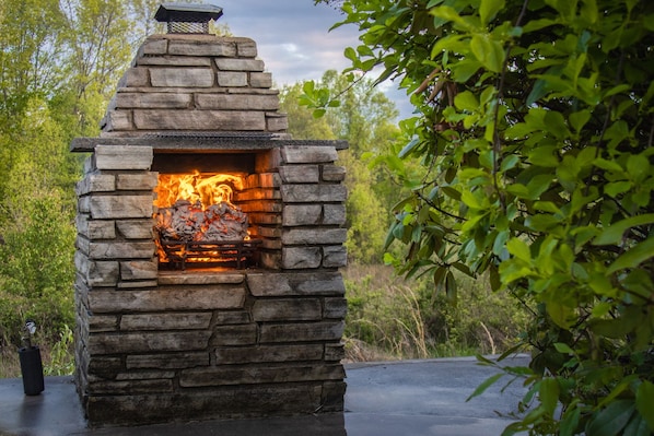 A stone-built outdoor fireplace with a fire burning inside, surrounded by greenery.