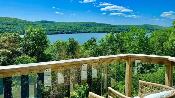 A wooden deck with glass railings overlooking a lake surrounded by lush greenery and hills.