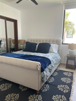 Une chambre à coucher comprenant un lit, des tables de chevet, un miroir et une fenêtre donnant sur des palmiers.