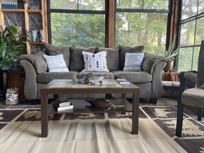 Relax in this inviting living room with a sofa, coffee table, and bookshelf, surrounded by lush greenery views.