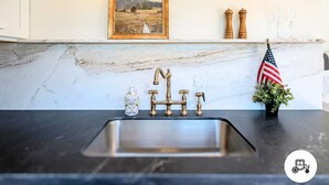 Una cocina con encimera de mármol, un lavamanos de acero inoxidable, un grifo de estilo vintage, un cuadro enmarco con paisaje, saleros y pimenteros de madera, una bandera estadounidense y una pequeña planta en maceta.