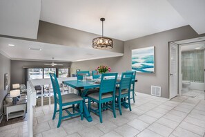 A spacious dining area with a large table and chairs, a ceiling fan, and a view of the ocean through large windows. - Across the Street to Siesta Key Beach, Tennis. Pickleball, Park, Heated Pool | Waterfront Balcony! (Siesta Key)