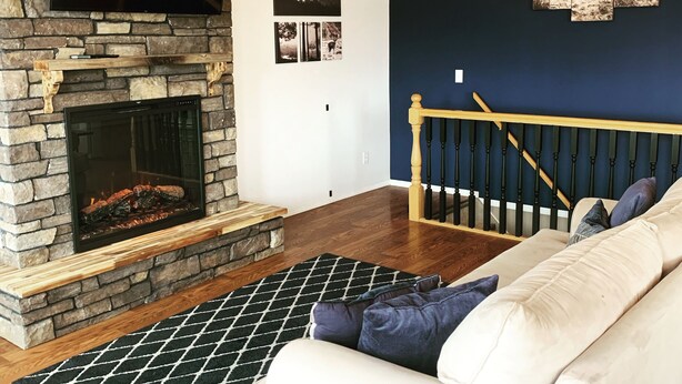 A living room with a stone fireplace, a flat-screen TV, a ceiling fan, and a staircase with a wooden railing.
