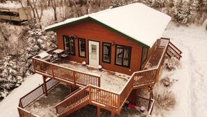 A cabin with a wooden deck and a covered porch, surrounded by snow.
