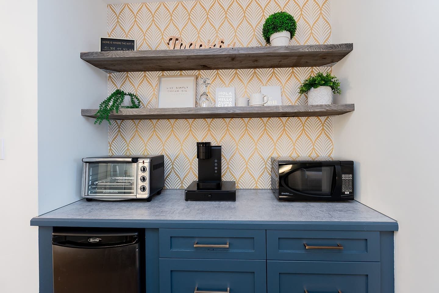 Una cocina con tostadora, cafetera y microondas sobre una encimera azul, y repisas decorativas arriba.