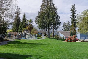 Zona residencial con un césped cuidado, un parque infantil con tobogán y un gran árbol en el centro.