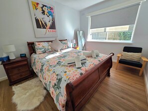 A bedroom with a wooden bed, floral bedding, a nightstand with a lamp, a chair, and a window with a view. - Luxurious 2BR: Near Showgrounds & Racecourse! (Moonee Ponds)