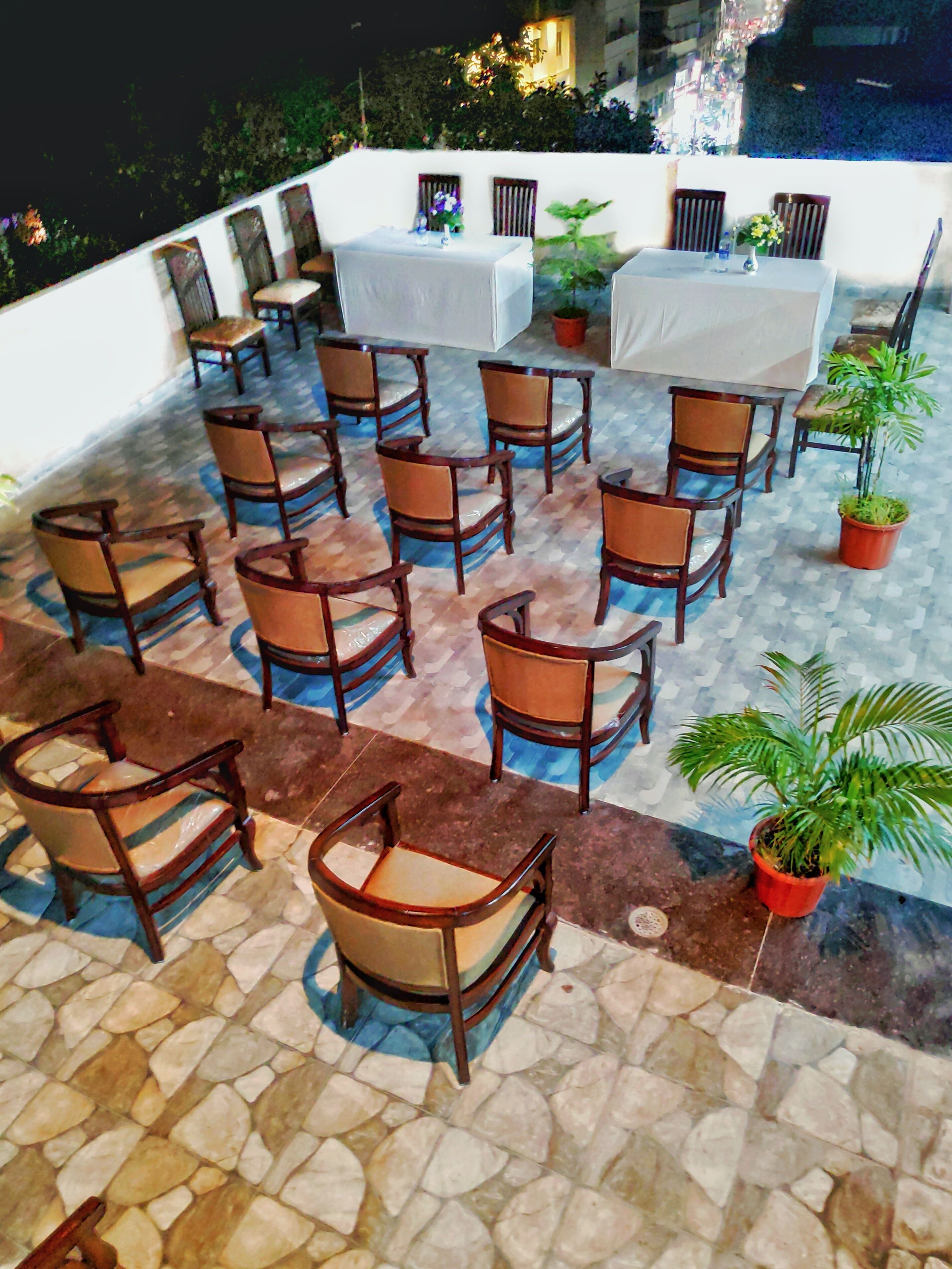 A rooftop terrace with multiple wooden chairs and tables, potted plants, and a white wall in the background.