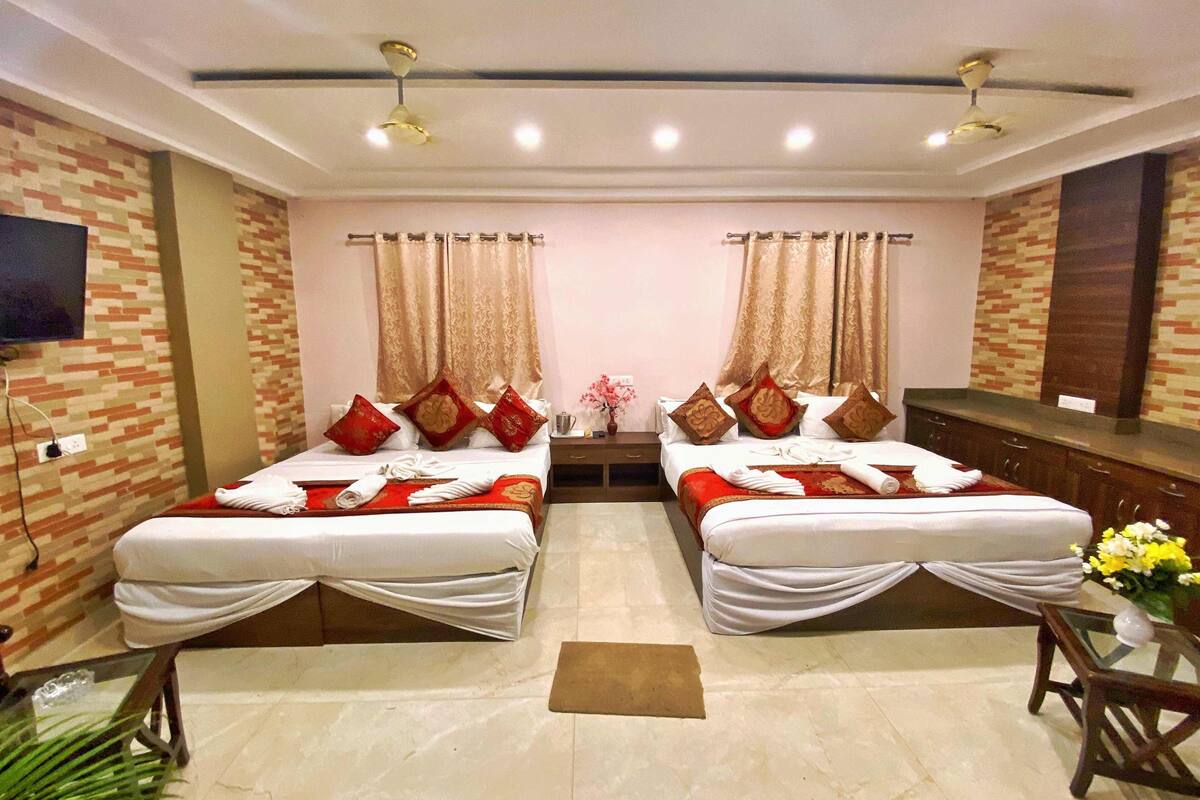 Habitación de hotel con dos camas, cada una con almohadas rojas y doradas, una mesita con un florero y un televisor en la pared.