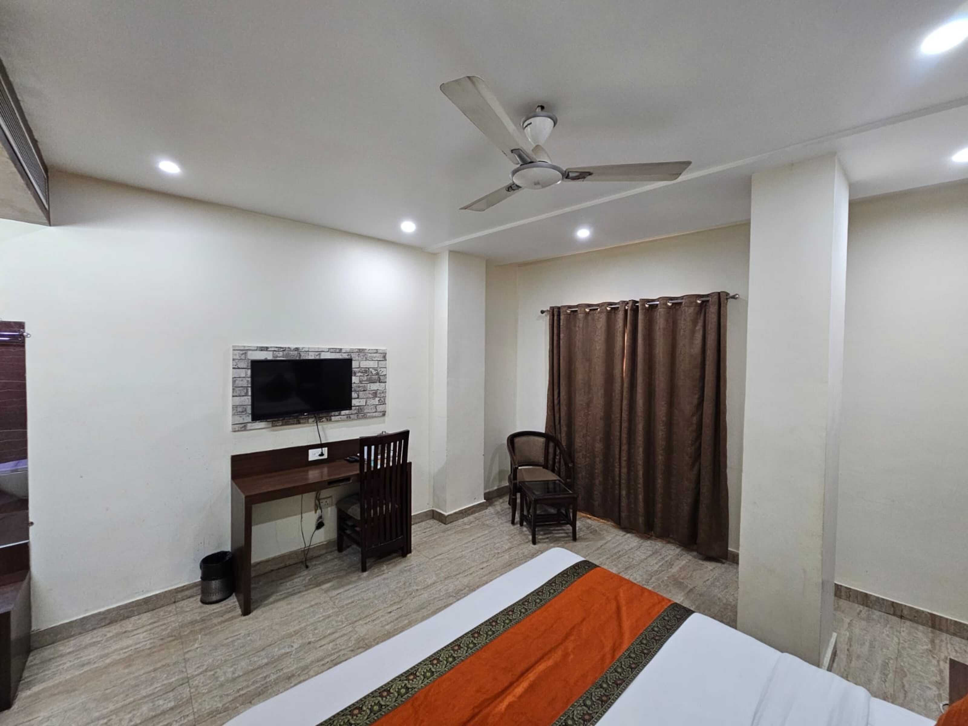 Una habitación de hotel con una cama, un escritorio con silla, un televisor montado en la pared, un ventilador de techo y una ventana con cortinas.