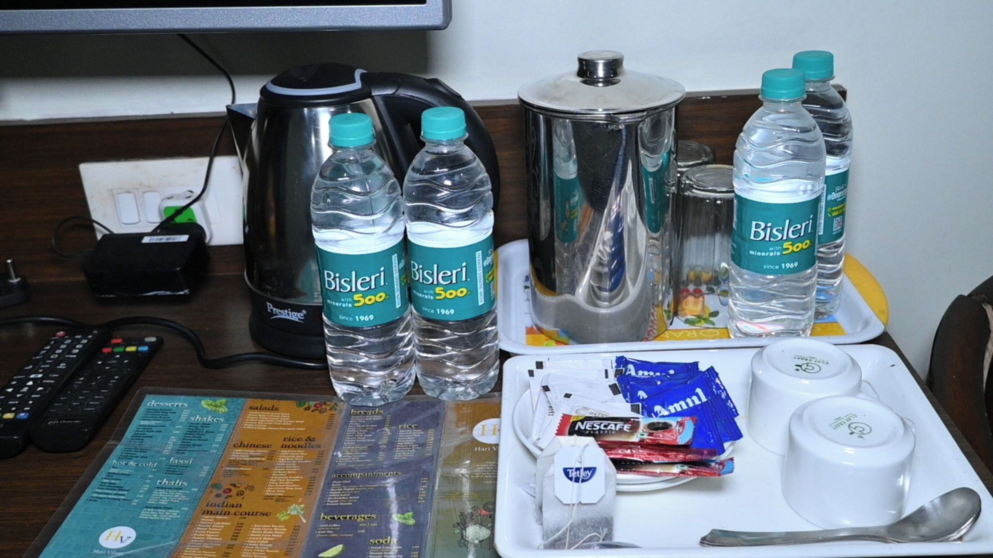 Camera d'albergo con un tavolo imbandito con bottiglie d'acqua Bisleri, un pacchetto snack Nescafe, un vassoio con una tazza e un cucchiaino, e un menu con diverse opzioni gastronomiche.