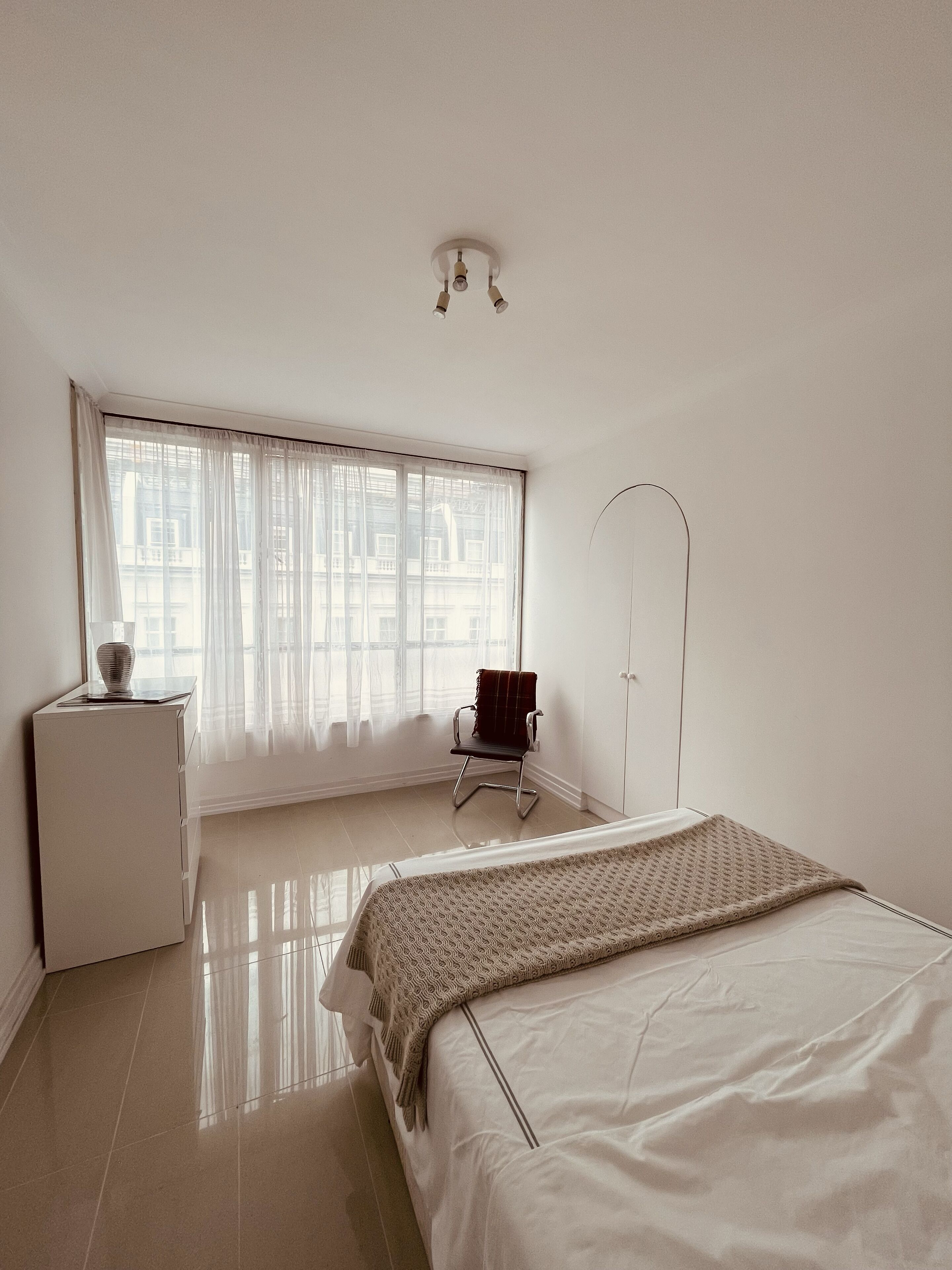 A minimalist bedroom with a bed, a chair, a desk, and a window with sheer curtains.