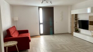 Un salón amplio con un sofá rojo, una mesita redonda, un televisor montado en la pared y un suelo de madera con diseño de espiga.