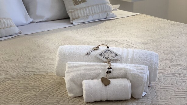 Una cama bien hecha con sábanas blancas y almohadas decorativas, una mesita de noche con lámpara, y un aplique en la pared.