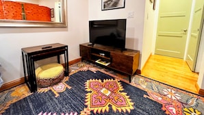Una habitación con una televisión sobre un soporte de madera, un espejo en la pared, una alfombra estampada y una puerta.
