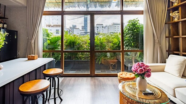 Un interior moderno con un gran ventanal, piso de madera, un sofá blanco, una mesa de centro de mimbre y una zona de bar con taburetes.