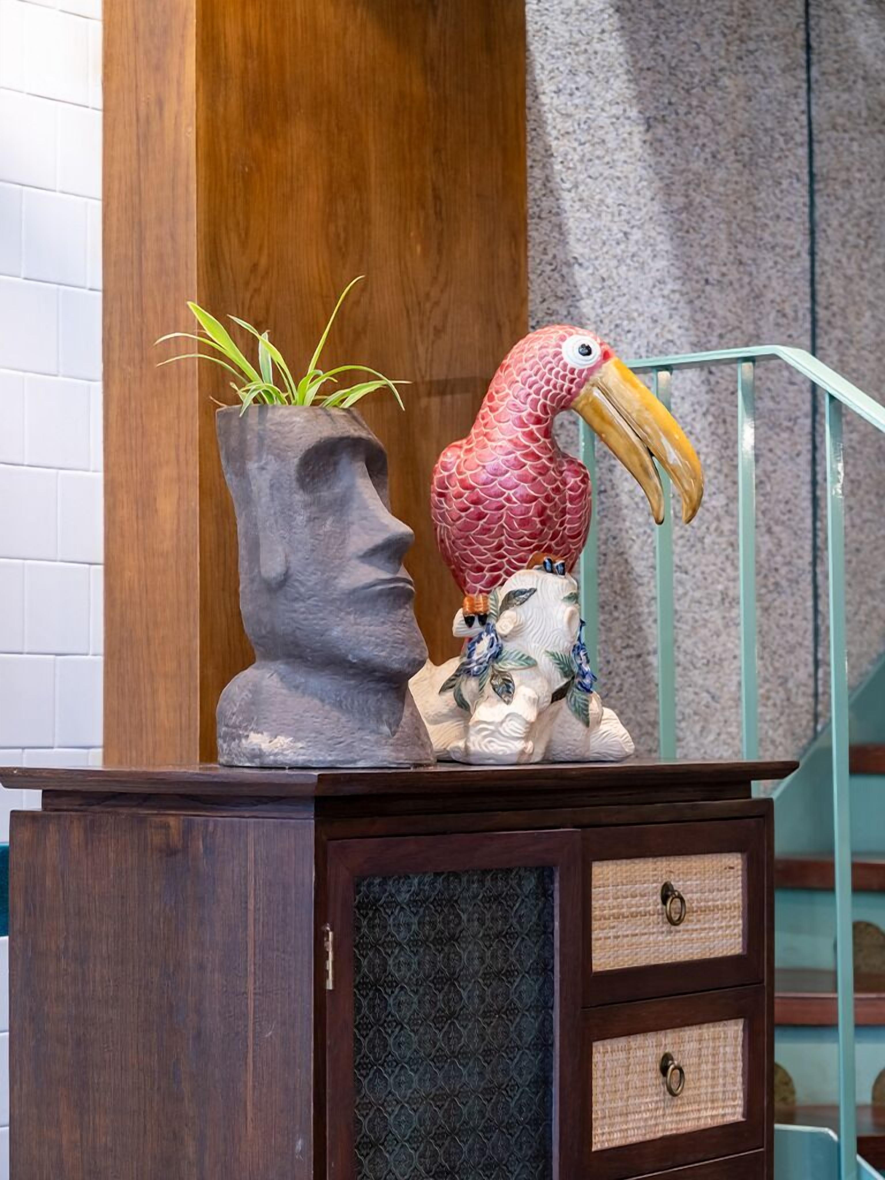 Un meuble en bois avec des tiroirs, une sculpture représentant un visage en pierre, une plante en pot et une figurine de perroquet.