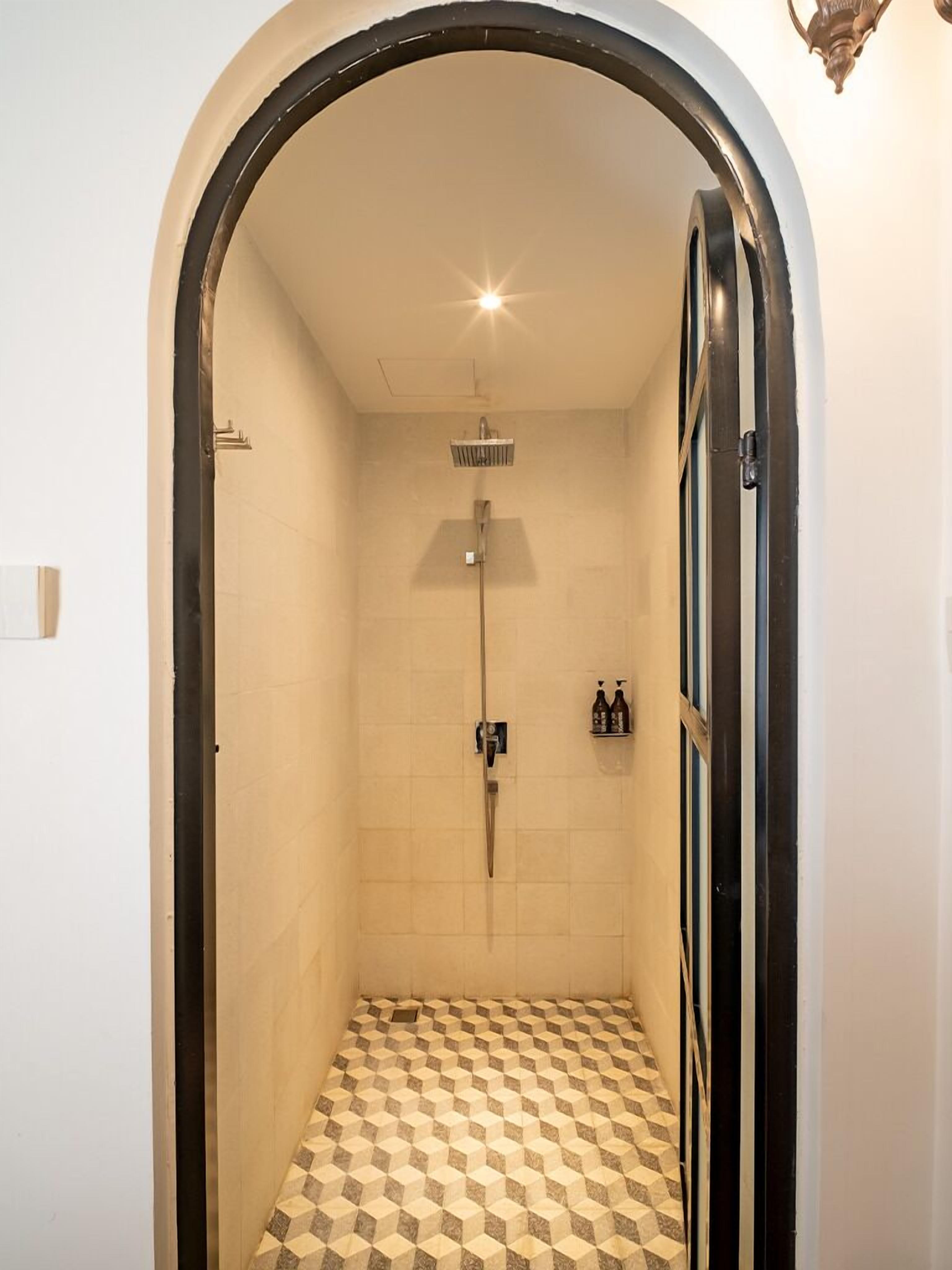 Un baño pequeño y cerrado con piso a cuadros, un cabezal de ducha y un estante en la pared con botellas.
