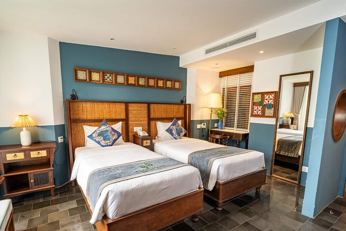 Een hotelkamer met twee bedden, een houten hoofdbord, een nachtkastje, een lamp, een spiegel en een raam met jaloezieën.