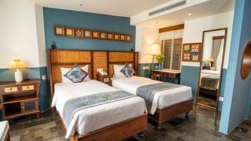 A hotel room with two beds, a wooden headboard, a nightstand, a lamp, a mirror, and a window with blinds.