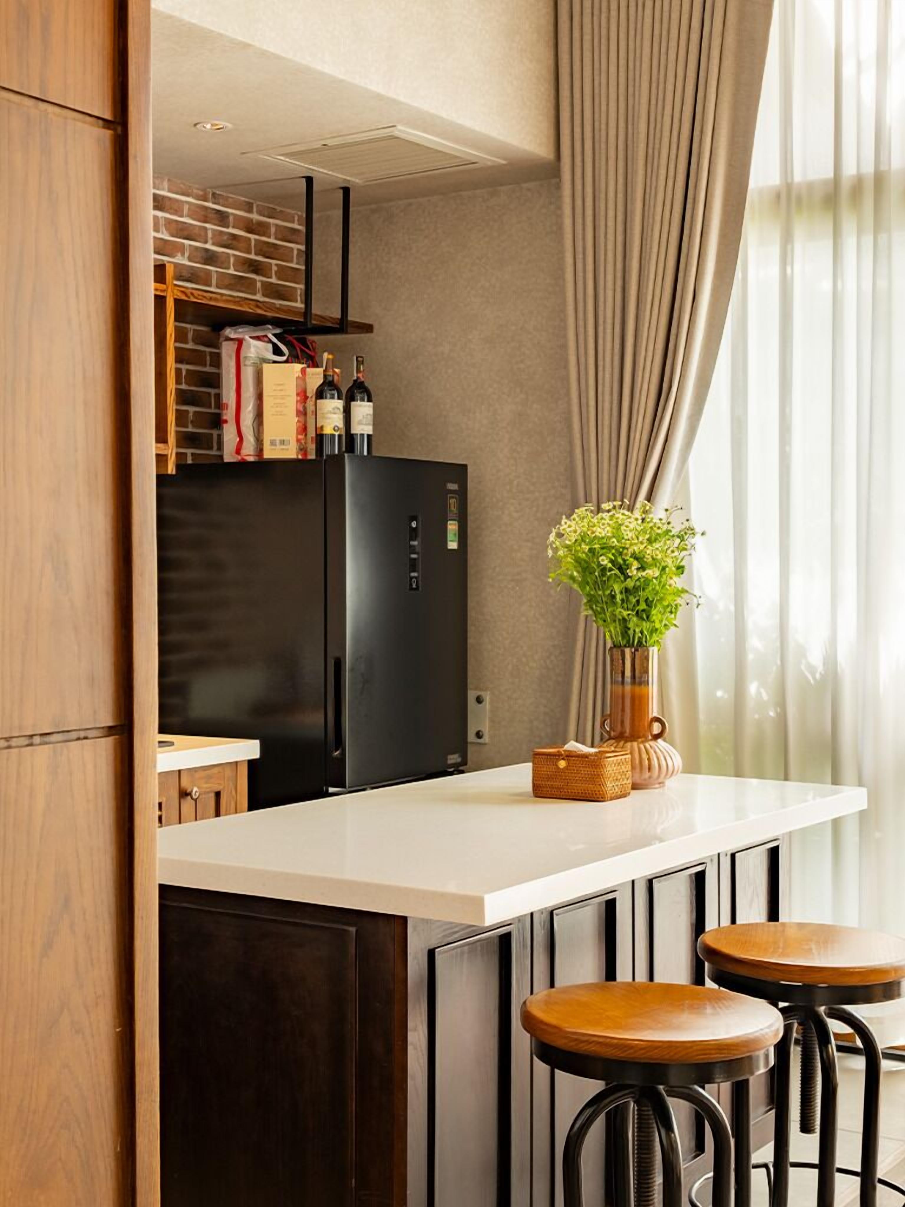 Eine moderne Küche mit einem schwarzen Kühlschrank, Holzschränken und einer weißen Arbeitsplatte. Auf der Arbeitsplatte stehen zwei Hocker und ein Blumentopf.
