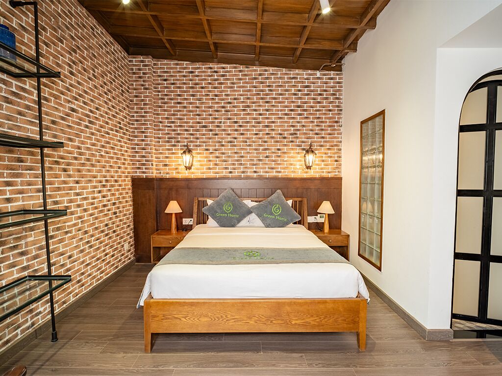 A hotel room with a brick wall, wooden ceiling, a bed with a wooden frame, two bedside tables with lamps, and a large window with a view.