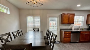 A dining area with a rectangular table and chairs, a kitchen with wooden cabinets and a dishwasher, and a ceiling light fixture. - 4 Bedroom House with Game Room (Lexington)