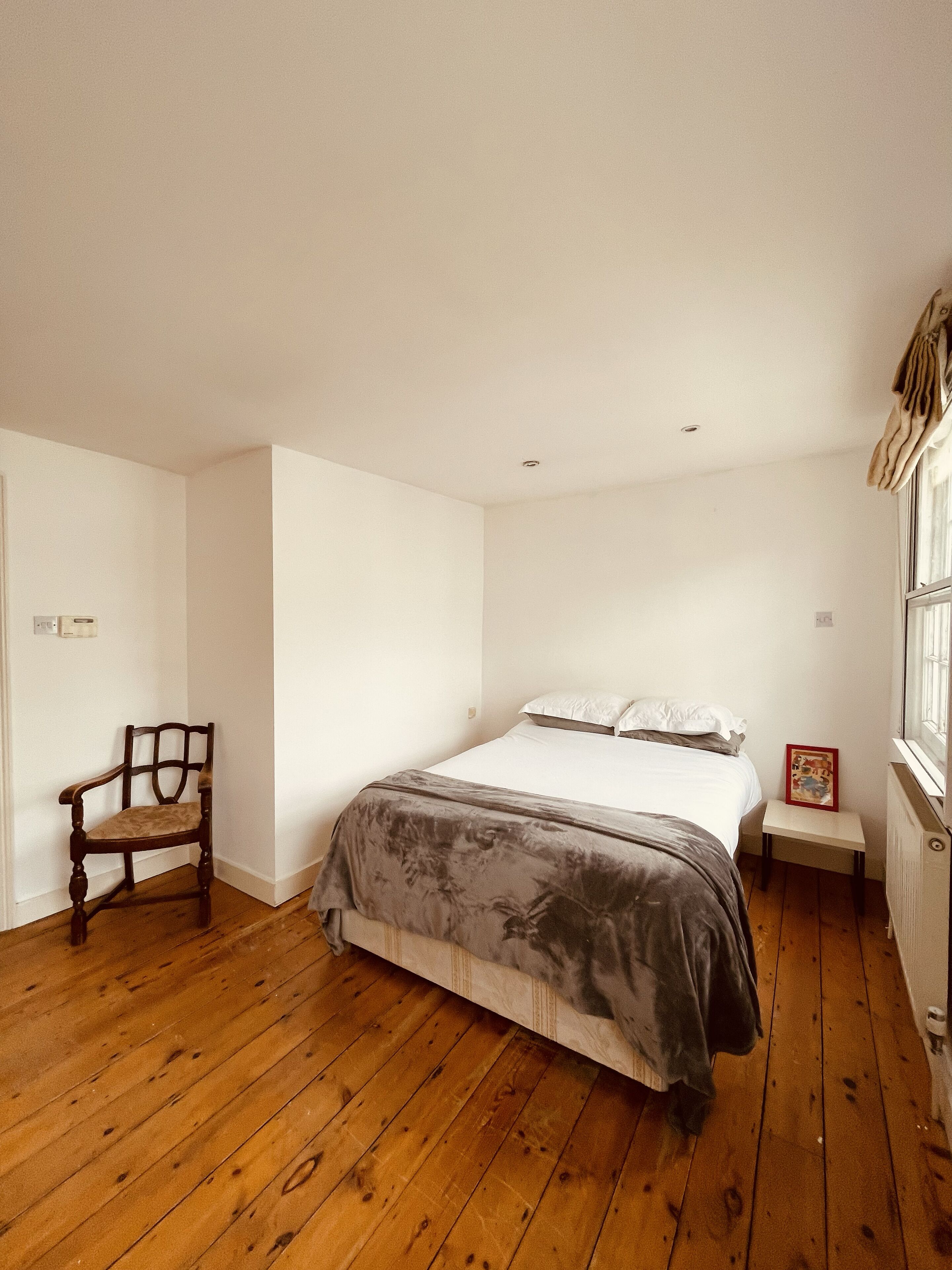 A bedroom with a wooden floor, a bed with white bedding, a chair, and a small table with a picture frame.