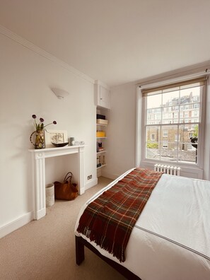 A bedroom with a bed, a white console table with a vase and picture, a window with a view of buildings, and a radiator.