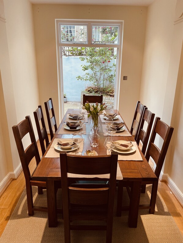 A dining table set for six with wooden chairs, a vase of flowers, and a view of a garden through a window.