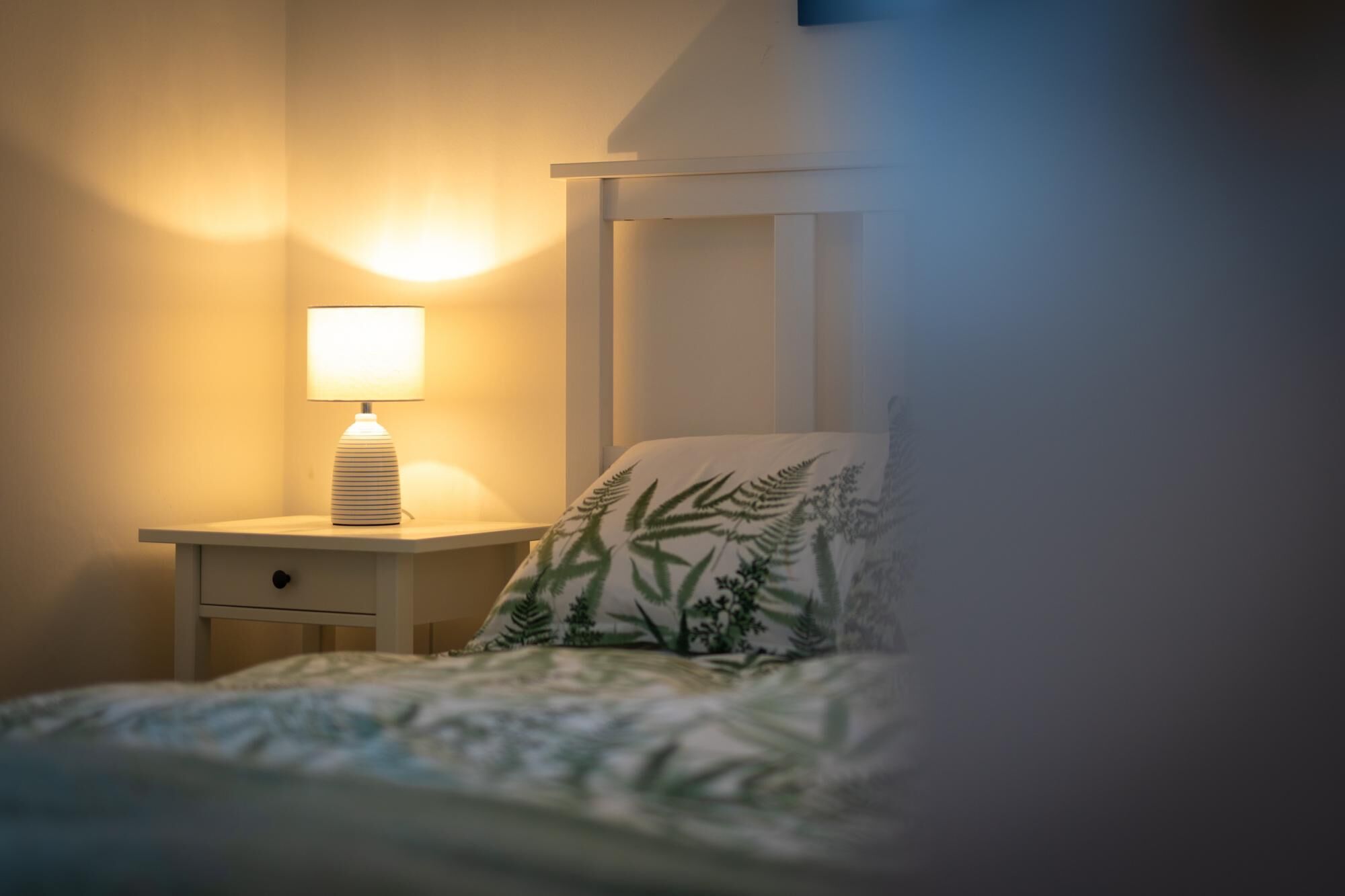 A bedroom with a nightstand, a lamp, a bed with a leaf-patterned pillow, and a white headboard.