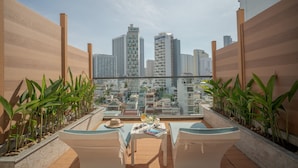Terraza en la azotea con vistas a un paisaje urbano, que incluye una mesa con una bebida, un sombrero y gafas de sol.