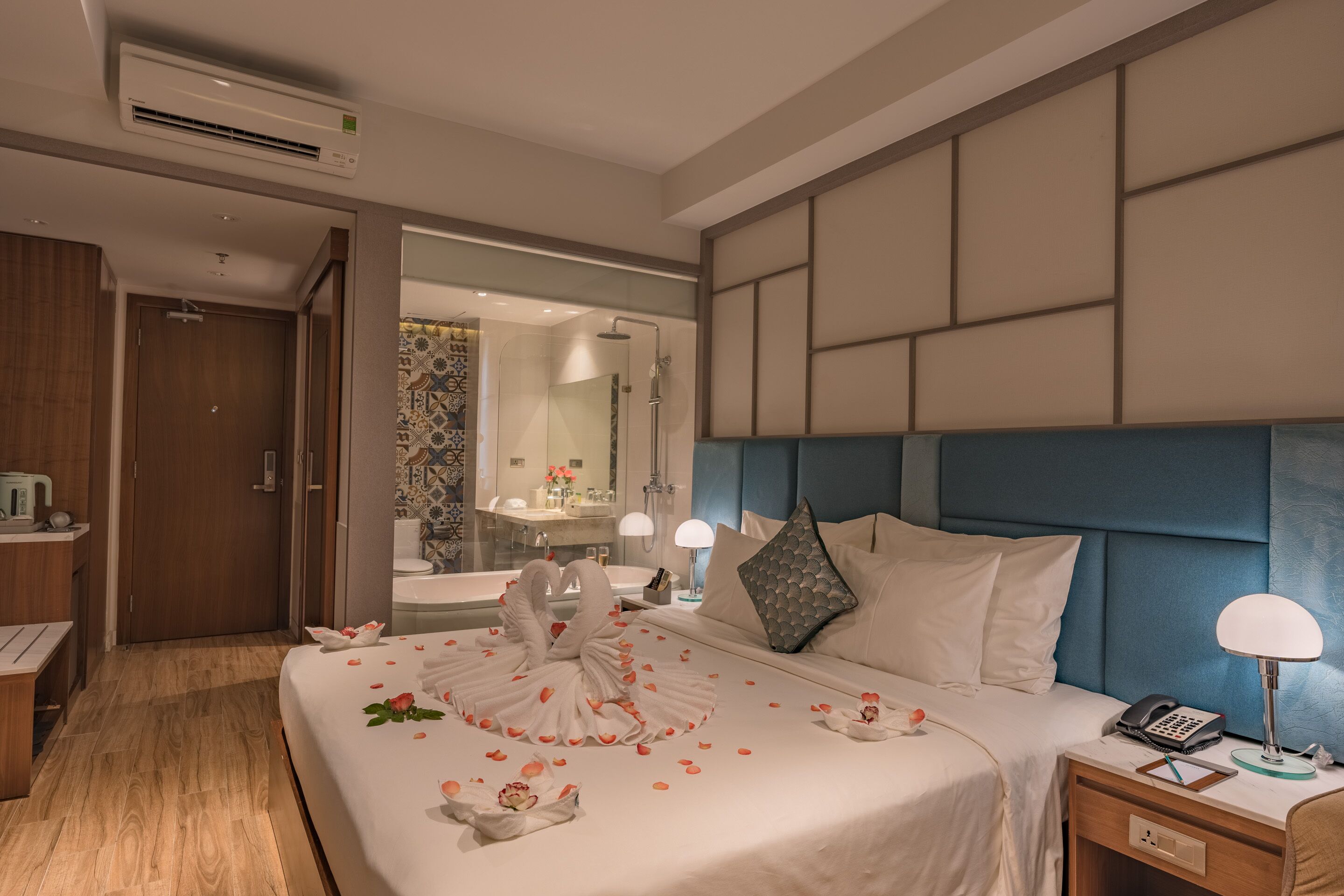 A hotel room with a large bed, a bedside table with a lamp, a mirror, and a bathroom visible through an open door.