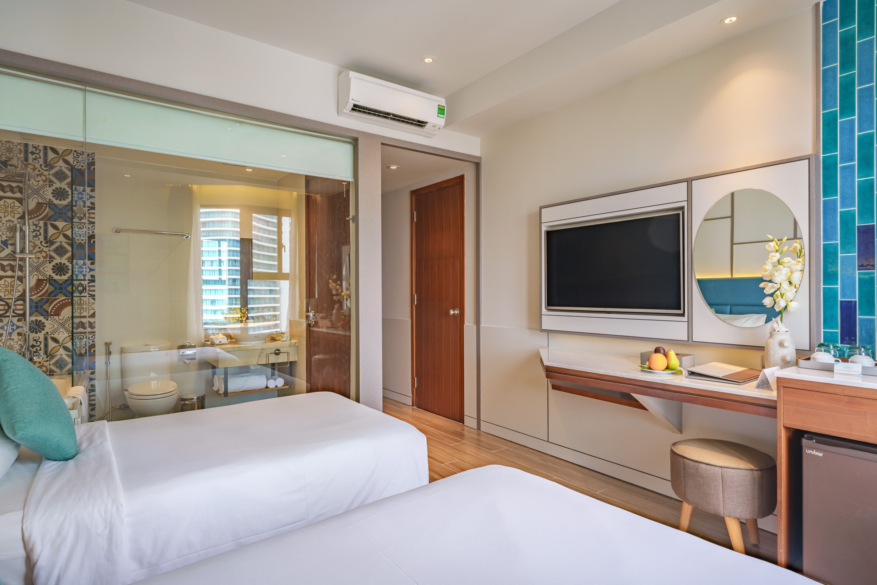 A modern hotel room with two beds, a TV, a minibar, and a bathroom with a glass shower enclosure.