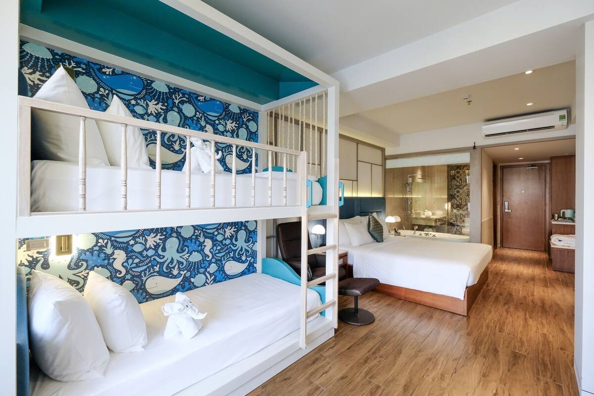 Habitación de hotel con literas, una cama individual, baño con ducha de vidrio y suelo de madera.