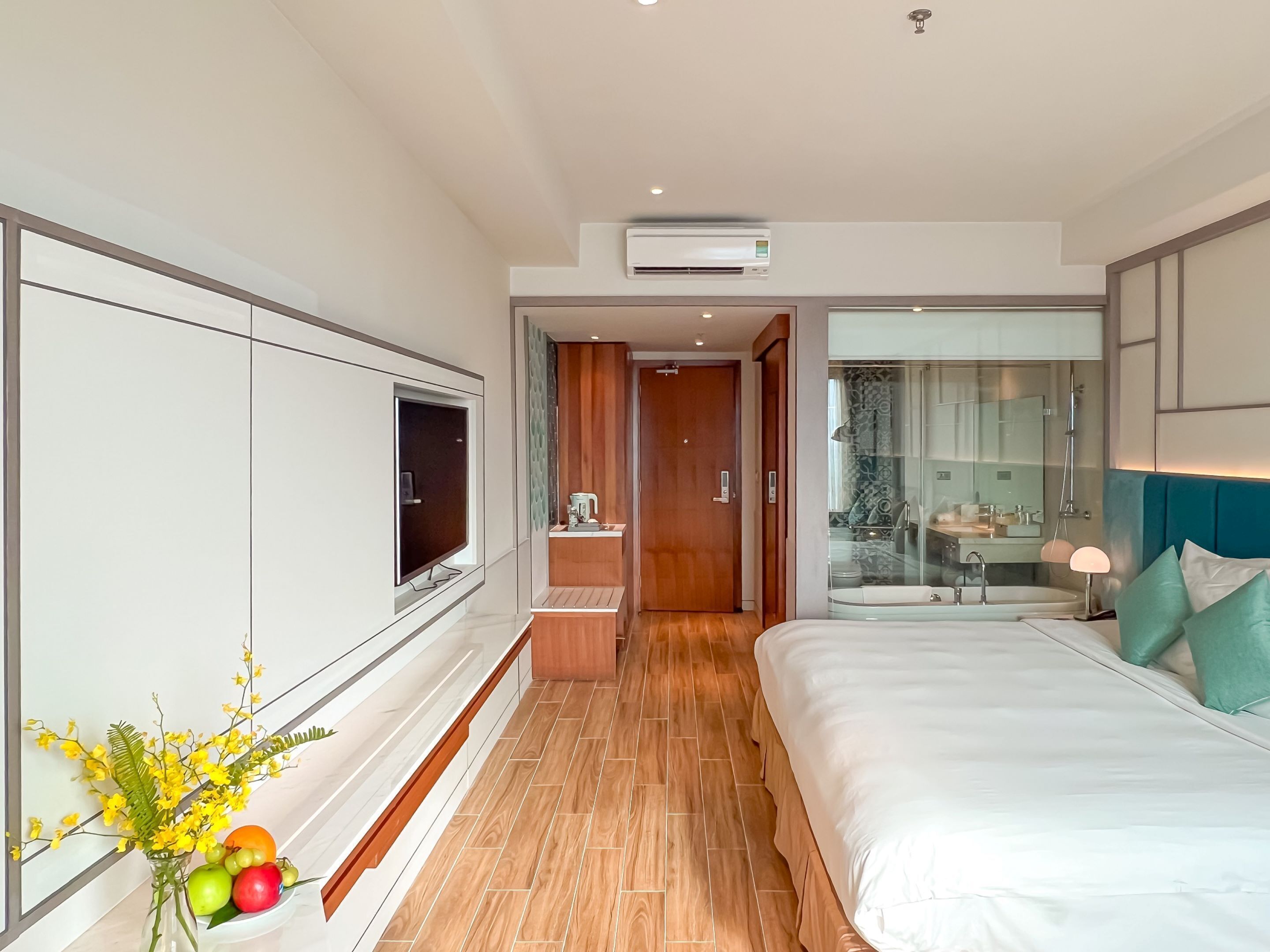 A modern hotel room with a large bed, a TV, a wooden floor, and a bathroom with a glass shower.