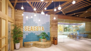 Un interior moderno con un llamativo letrero 'SEAESTA', vigas de madera en el techo, luces colgantes, una zona de estar con una planta y un panel de vidrio.