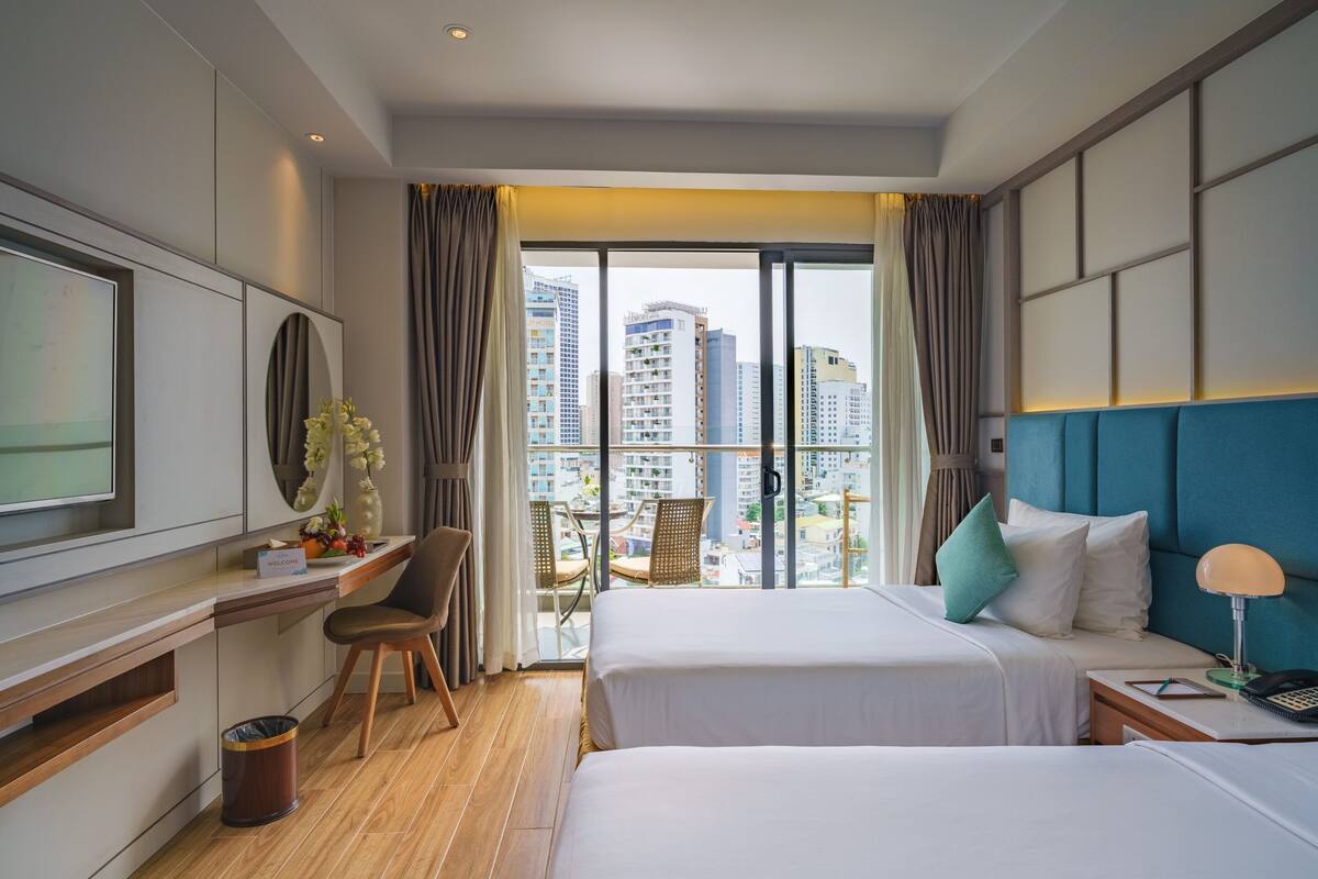 Habitación de hotel con dos camas, un escritorio, televisión y balcón con vistas a la ciudad.