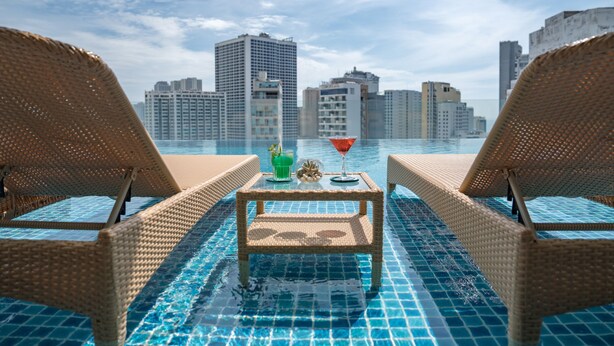 Vista de la piscina con sillones de mimbre, una mesa con una bebida y un paisaje urbano al fondo.
