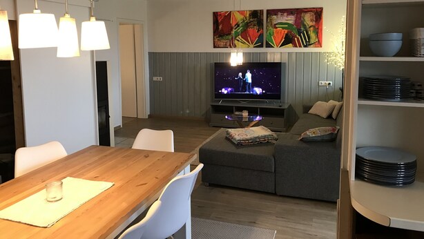 A modern living room with a wooden dining table, white chairs, a grey sofa, a flat-screen TV, and a clock on the wall.