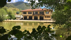 Edificio de dos pisos con tejado de tejas rojas, un balcón y un garaje. Se encuentra junto a una masa de agua tranquila que refleja el edificio.