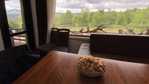 Una habitación con vista a un lago y árboles, una mesa de madera con una cesta de aperitivos y una ventana con cortinas.