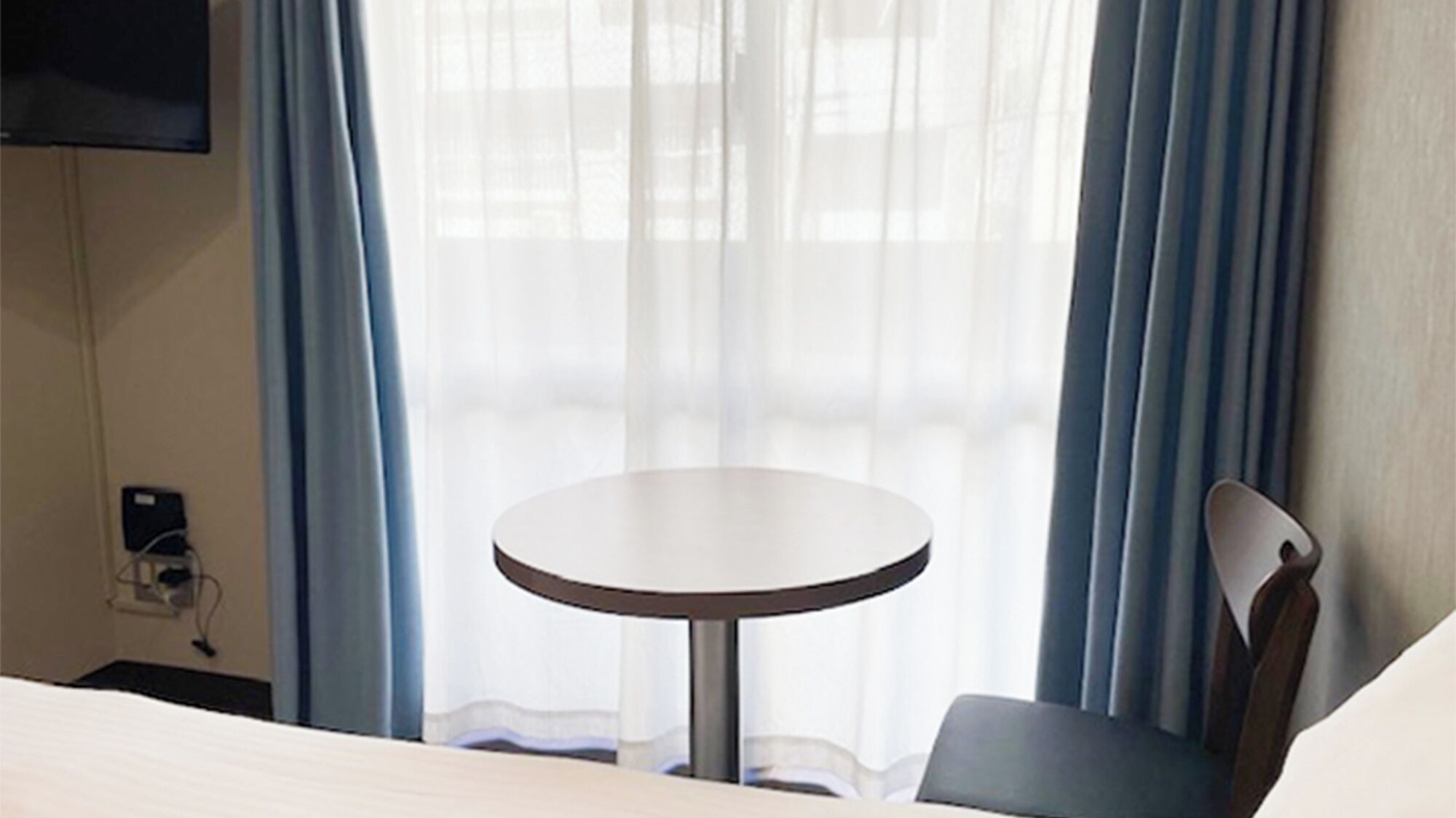 A hotel room with a round table, a chair, a bed, and a TV mounted on the wall.