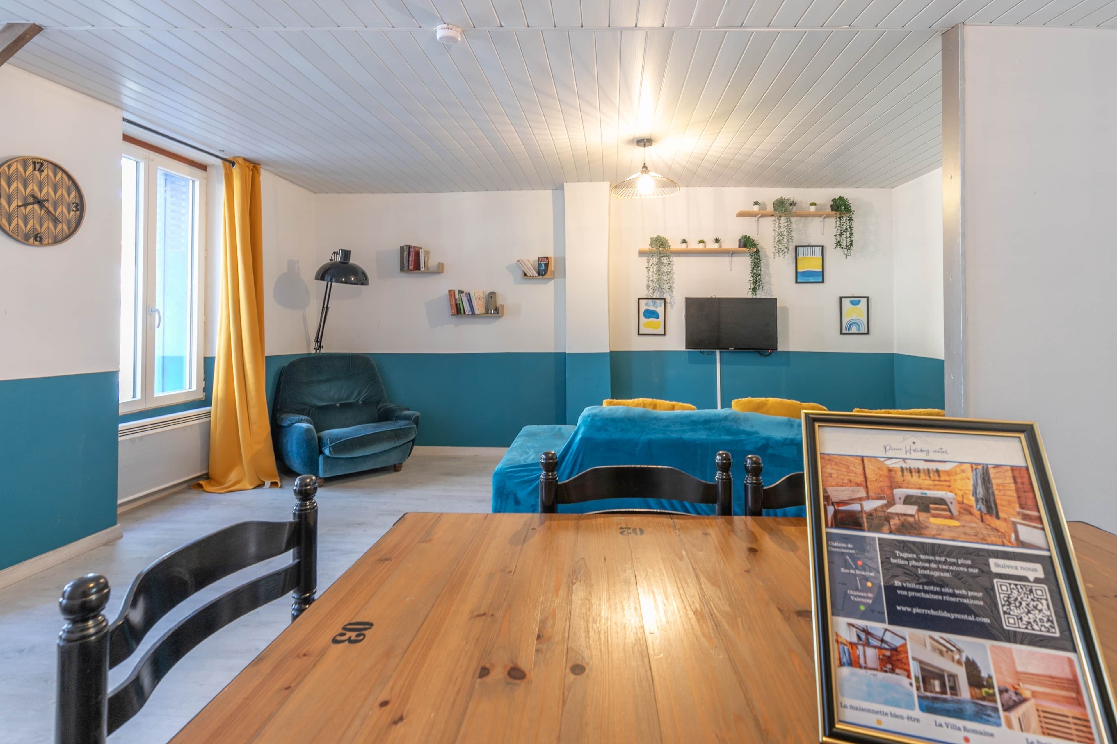 Ein Zimmer mit einem Holztisch, einem blauen Sofa, einem blauen Sessel, einem Fernseher und einem gerahmten Foto an der Wand.