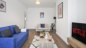 A modern living room with a blue sofa, a beige sofa, a coffee table with decorative items, a TV displaying 'NETFLIX', and a framed artwork on the wall. - BIG House Free Parking Walking Distance to Kensal Rise Station (London)