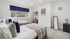 A bedroom with two beds, a window with a blue blind, a clock, a framed artwork, and a dresser with a lamp. - BIG House Free Parking Walking Distance to Kensal Rise Station (London)